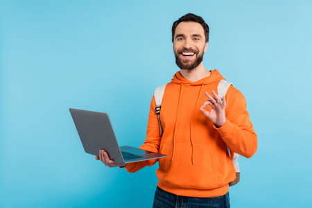 happy bearded student holding laptop and showing okay sign isolated on blueの写真素材