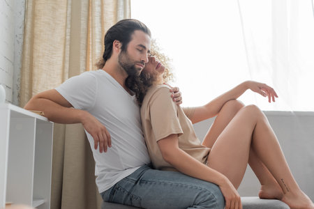 young woman with love me more tattoo lettering on leg sitting with bearded boyfriend on bed benchの写真素材