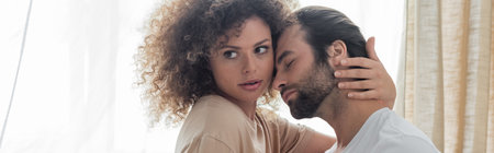 curly woman in beige t-shirt hugging bearded boyfriend in bedroom, banner