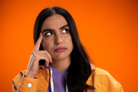 Portrait of pensive woman holding finger near eyebrow isolated on orange