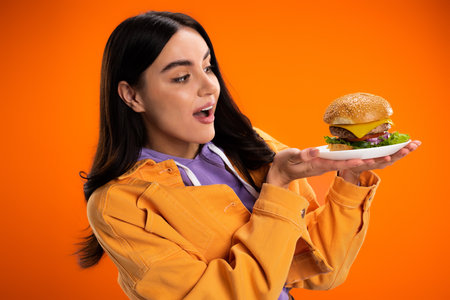 amazed woman in stylish jacket looking at tasty burger with cheese and meat isolated on orangeの写真素材