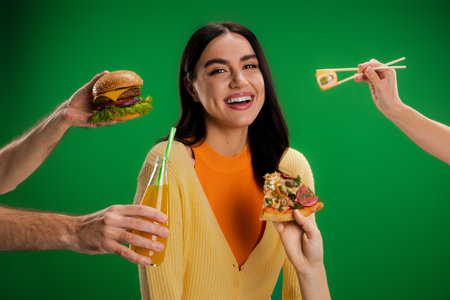 happy young woman looking at camera near people proposing different food isolated on green