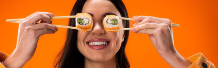cheerful brunette woman covering eyes with fresh sushi rolls isolated on orange, bannerの写真素材