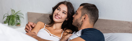 Man in pajama hugging smiling girlfriend on bed, banner