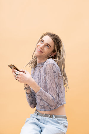 Smiling queer person in crop top with snakeskin print and jeans using smartphone isolated on yellow