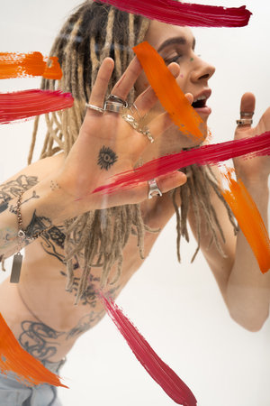 tattooed nonbinary person in silver rings touching glass with multicolored paint strokes on white...