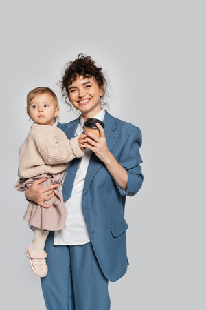 happy businesswoman in blue formal wear holding in arms toddler daughter and coffee to go isolated on greyの写真素材