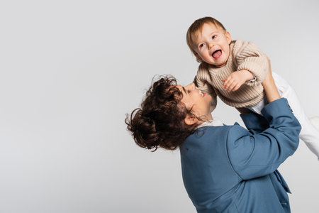 happy businesswoman in blue suit holding excited toddler girl in sweater isolated on grey