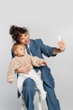 happy businesswoman in blue suit taking selfie with toddler daughter on chair isolated on grey