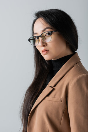 young asian businesswoman in glasses and blazer looking at camera isolated on grey