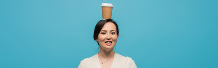 Smiling asian woman holding coffee to go on head isolated on blue, banner