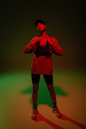 full length of young african american woman in baseball cap and sweatshirt adjusting vest while...