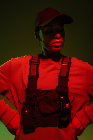 african american woman in sweatshirt and vest posing with hands on hips on green with red lightの写真素材
