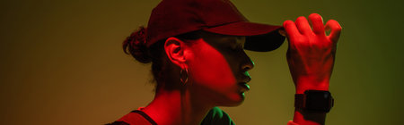 side view of african american woman in sweatshirt adjusting baseball cap on green with red light, bannerの写真素材