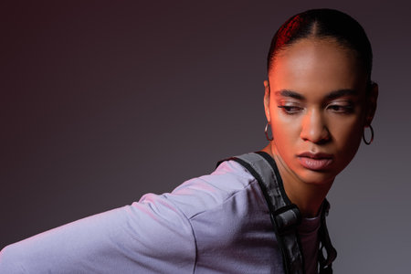 brunette african american woman in sweatshirt and vest looking at camera on grey and purple...