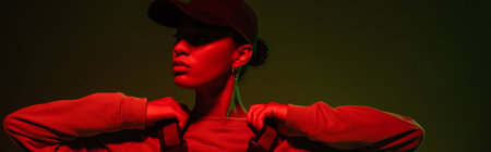 african american woman in sweatshirt and baseball cap posing on green with red light, bannerの写真素材