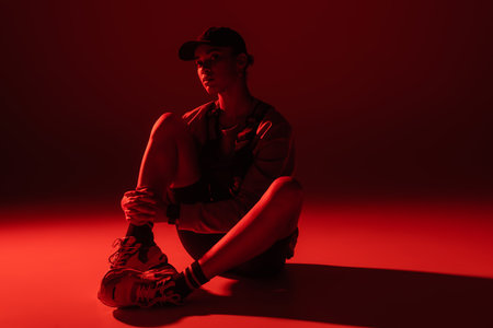 full length of young african american woman in baseball cap and sweatshirt sitting on dark red backgroundの写真素材