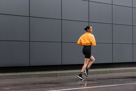 full length of brunette african american sportswoman in bike shorts and yellow puffer jacket jogging outsideの写真素材