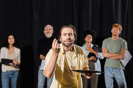 emotional man with clipboard looking at camera and gesturing during rehearsal near multiethnic actors and producer on blurred backgroundの写真素材
