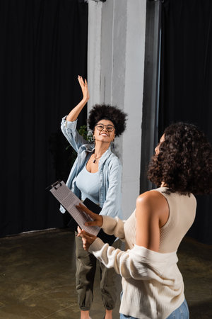 smiling african american actress in eyeglasses posing with raised hand near multiracial woman with screenplayの写真素材