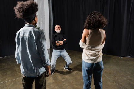 bearded teacher posing with crossed arms near multiethnic actresses in acting school. Translation of...