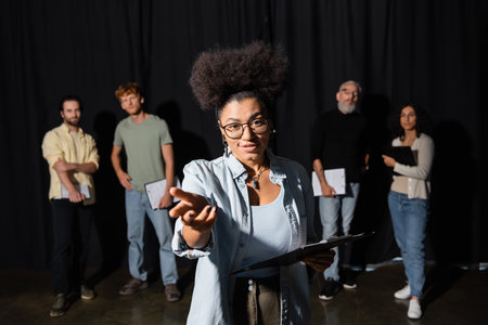 positive african american woman holding scenario and pointing with hand while rehearsing near blurred interracial students and art director in theaterの写真素材