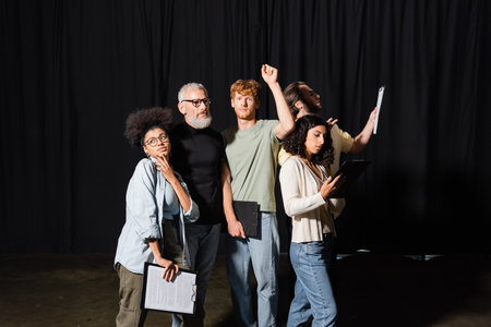 redhead man looking at camera while standing with raised hand near multiethnic actors and bearded producer in theaterの写真素材