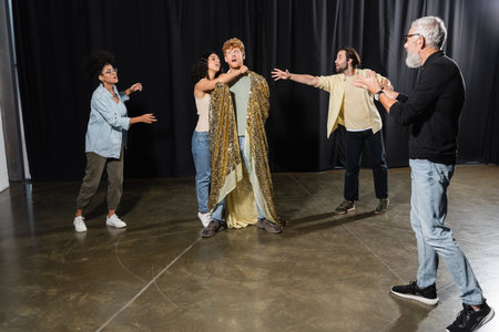 grey haired art director gesturing near interracial students rehearsing scene of king assassination in theaterの写真素材