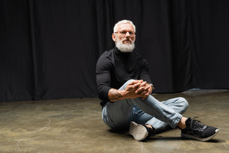 tattooed actor in eyeglasses and black turtleneck looking at camera and sitting on stage in theater. Translation of tattoo: om, shanti, peaceの写真素材