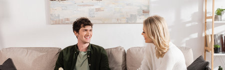 young and cheerful couple looking at each other in living room, banner