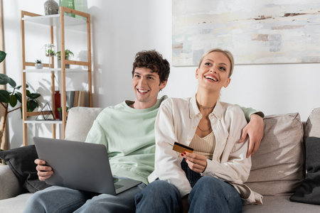 cheerful woman holding credit card near boyfriend with laptop doing online shoppingの写真素材