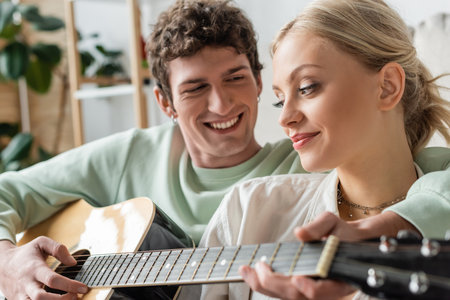 happy man playing acoustic guitar near blonde woman on blurred foregroundの写真素材