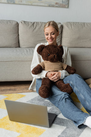 positive young woman in jeans holding teddy bear and watching movie on laptopの写真素材