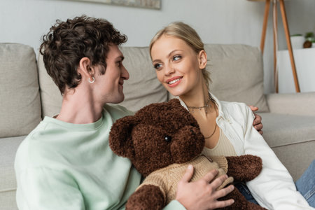 joyful young woman and happy man holding teddy bear in living roomの写真素材