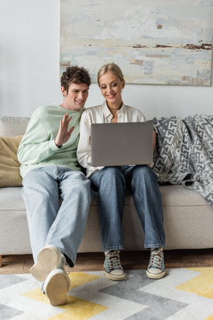 happy young man with curly hair waving hand near girlfriend during video call on laptopの写真素材