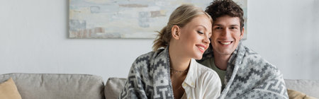 happy young man and woman covered in blanket smiling in living room, bannerの写真素材