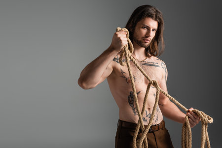 Young long haired model with tattoo on body holding rope isolated on greyの写真素材