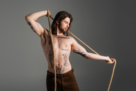 Muscular and tattooed man holding rope isolated on grey