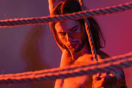 Shirtless and tattooed model posing near ropes on purple background with lightの写真素材