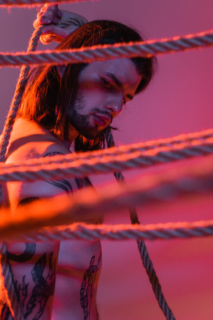 Tattooed man looking at blurred ropes on purple background with light
