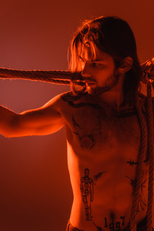 Long haired and tattooed man looking at rope isolated on redの写真素材
