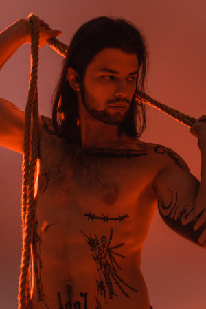 Young tattooed and shirtless model posing with rope isolated on red