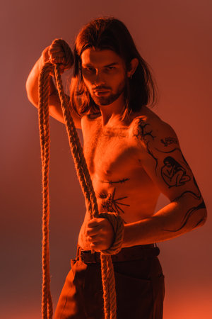 Long haired and tattooed model holding rope and looking at camera on red backgroundの写真素材