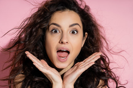 Portrait of shocked curly woman looking at camera on pink backgroundの写真素材