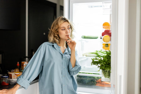 pensive young woman looking into open refrigerator in kitchenの写真素材