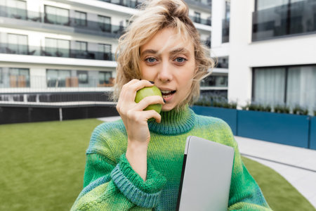 blonde woman in sweater holding laptop and eating green apple near hotel building in Barcelonaの写真素材