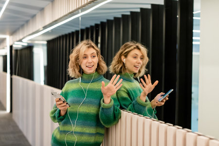 happy young woman listening music in wired earphones and holding smartphone while waving hand near...