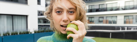 blonde woman in sweater eating green apple near hotel building, bannerの写真素材