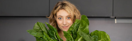 pierced young woman smiling and holding green cabbage leaves in kitchen, bannerの写真素材