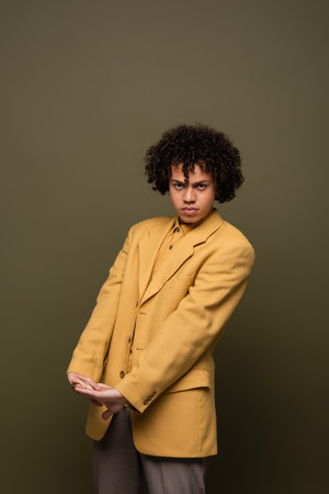 trendy african american man in yellow blazer looking at camera while posing with clenched hands on olive grey backgroundの写真素材